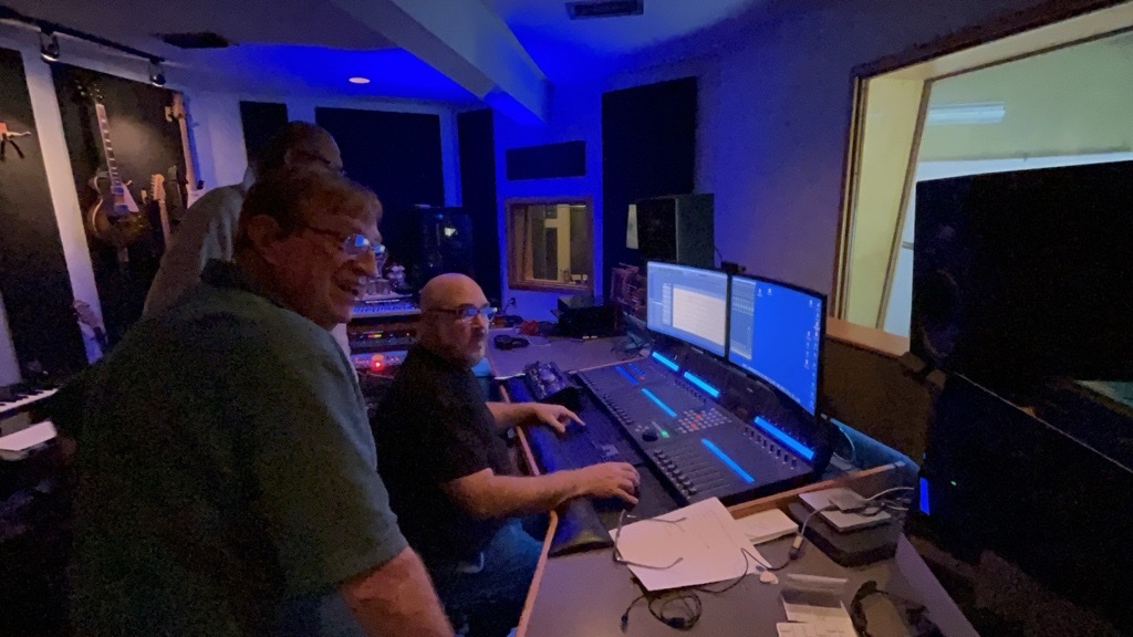 Two engineers work an audio console in a blue-lit control room, guitars racked on the wall behind.