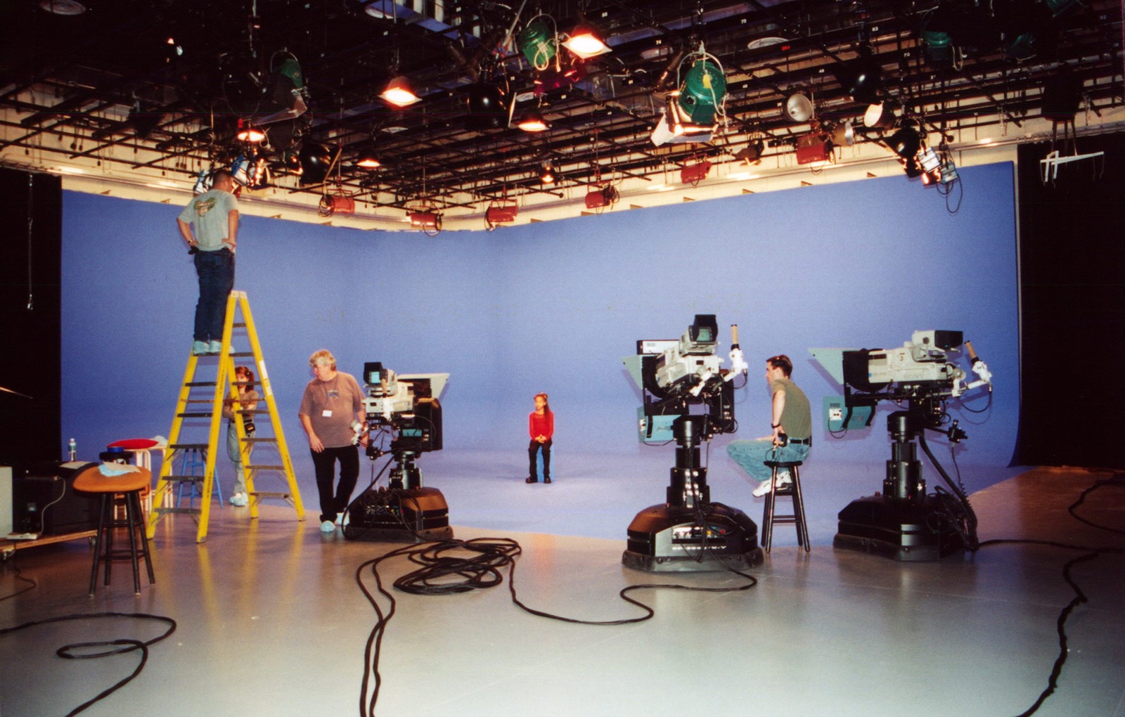 Three pedestal cameras and crew set up around a performer on a blue-screen stage.