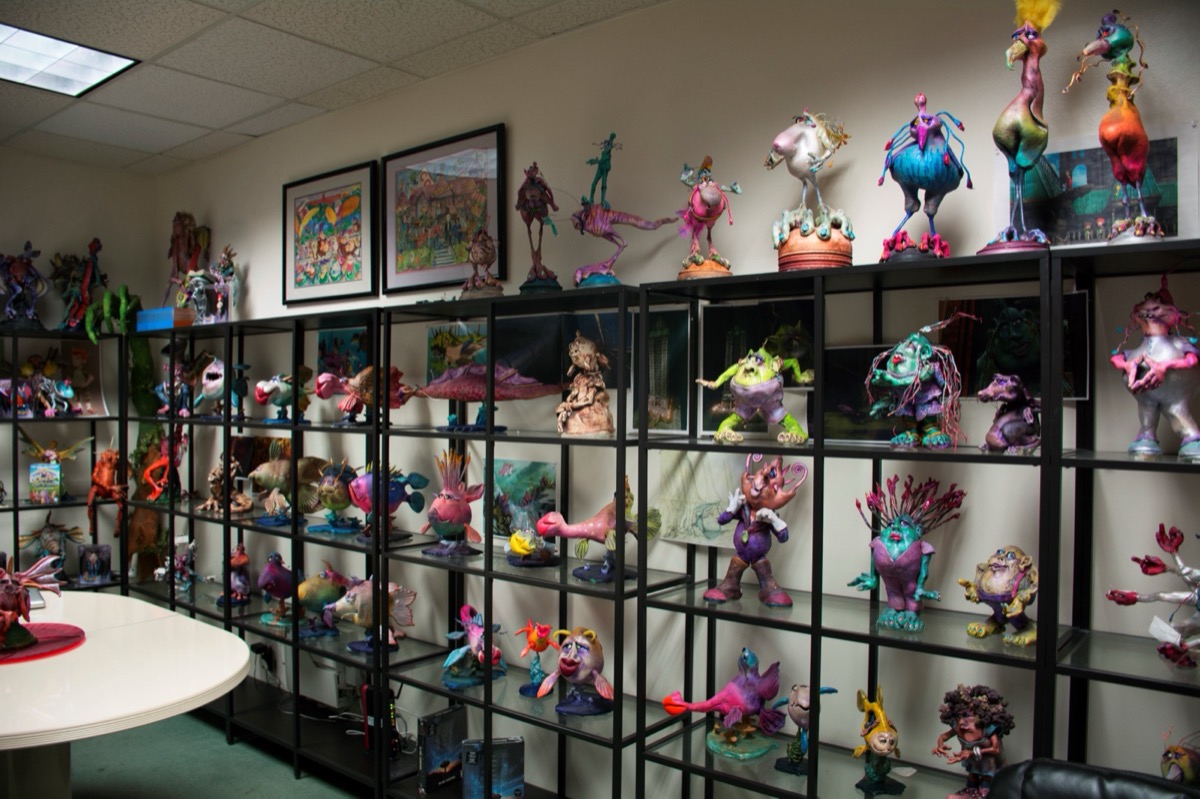A wall of black glass shelving in the TW3 character library, filled with dozens of hand-sculpted painted maquettes, framed concept paintings hung above and a conference table in the foreground.
