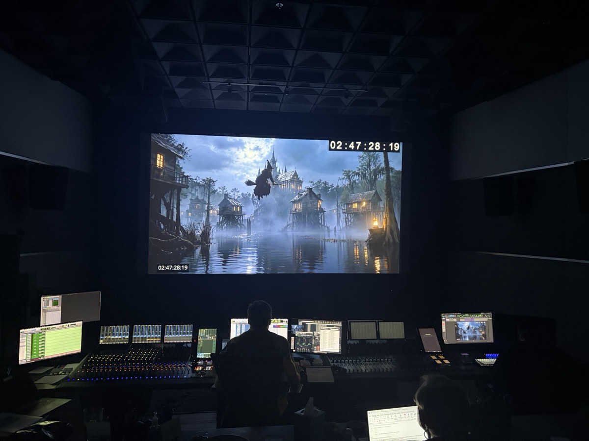 A mix engineer at the console of a dub stage, a wide shot from Merry The Christmas Witch on the screen above with timecode running.