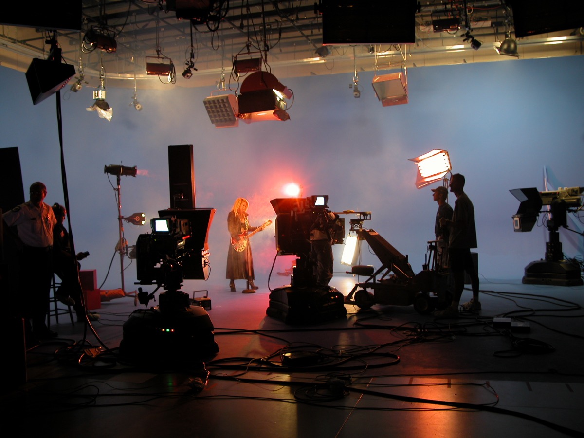 A guitarist performs under haze and red backlight while pedestal cameras, a jib arm and crew work in silhouette around her.