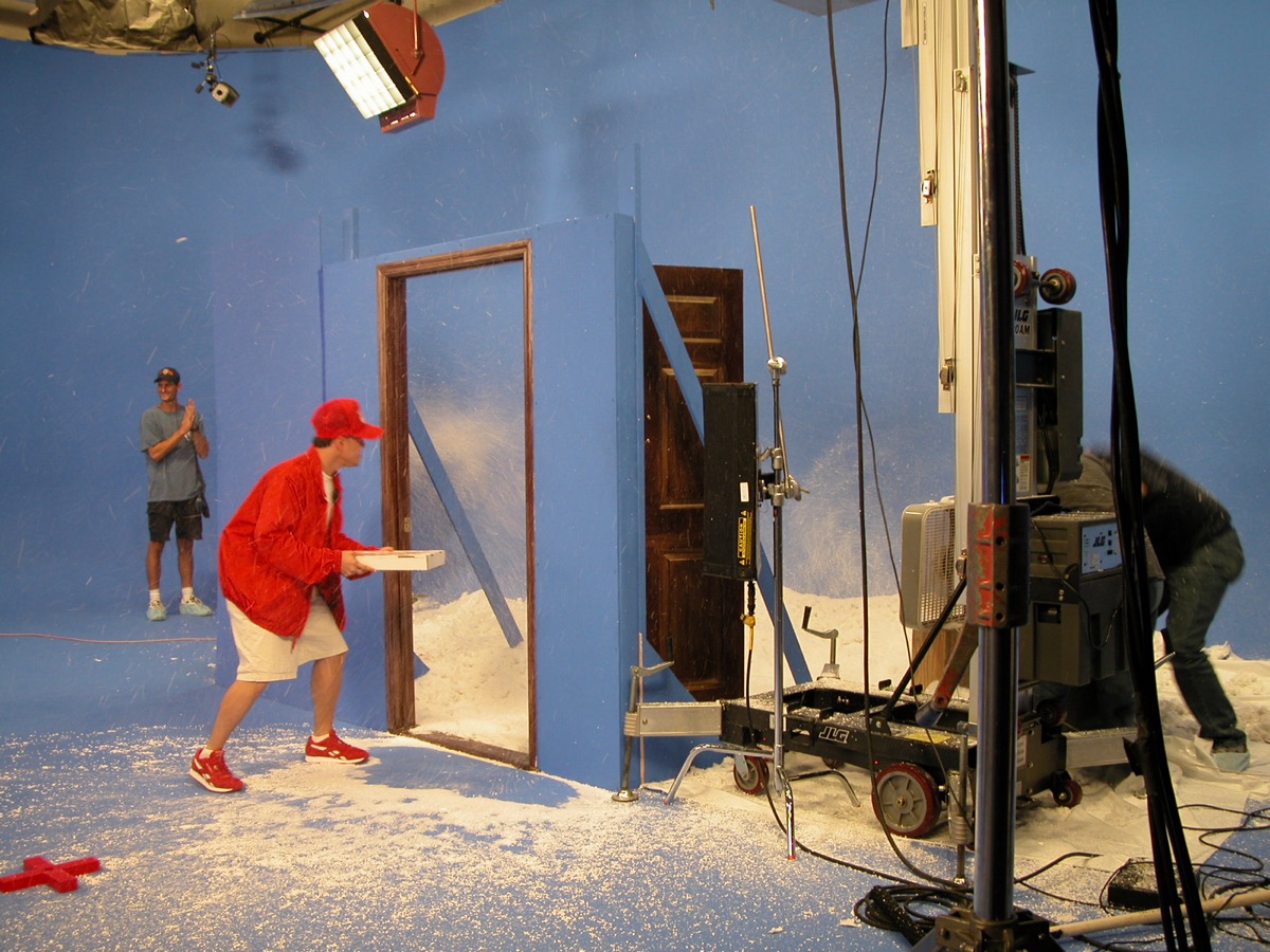 A performer in a red jacket carries a box through a doorway set buried in artificial snow on a blue-screen stage, crew and a lift alongside.