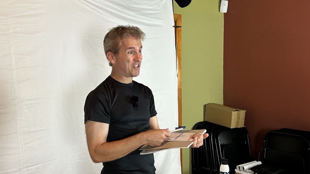 A presenter delivers to camera against a white backdrop, lavalier mic clipped on and notes in hand.