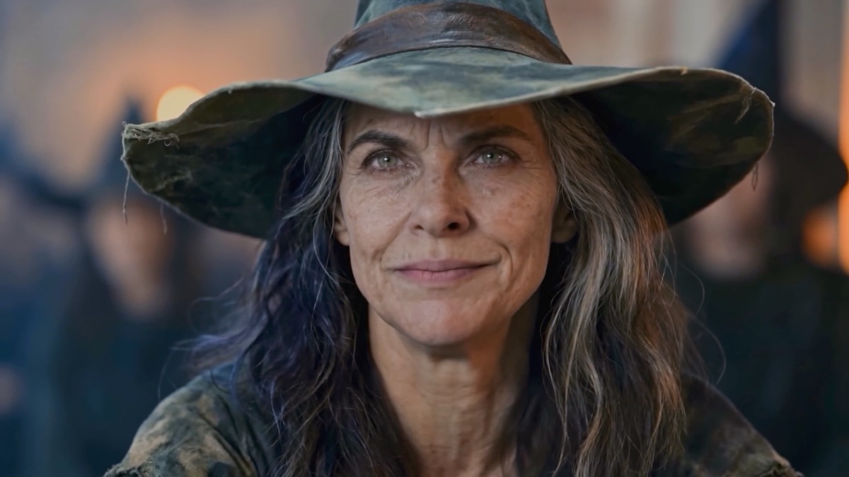 An elder witch in a weathered hat smiles warmly by firelight.