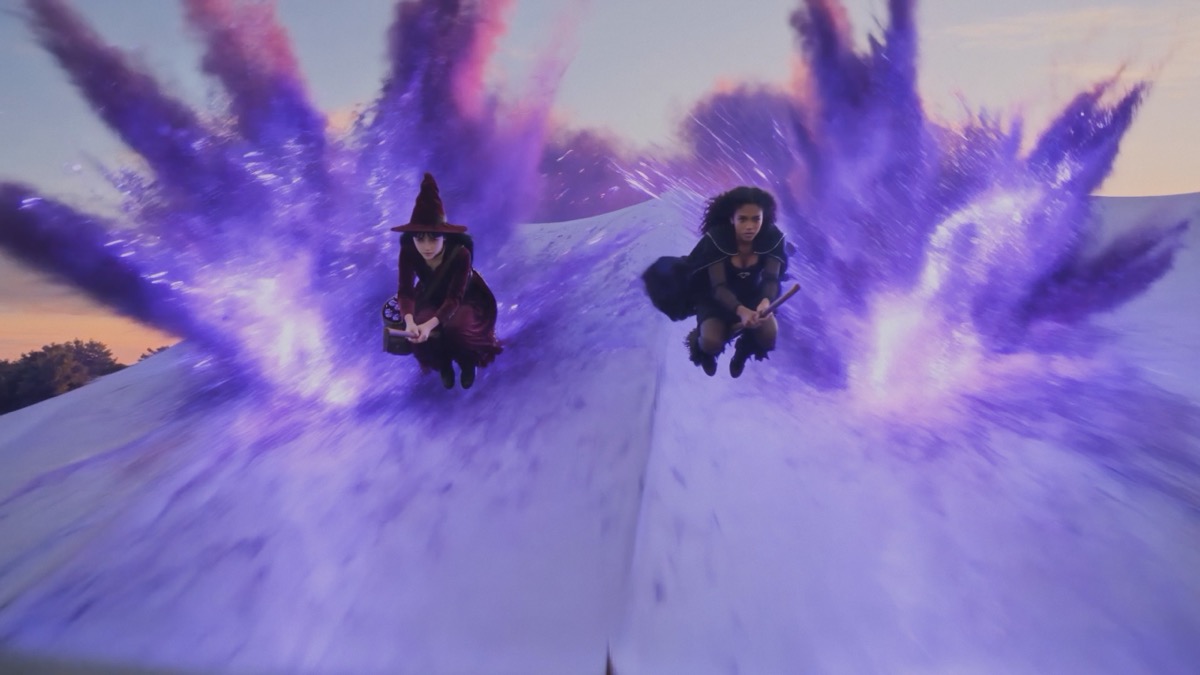 Two witches streak low over a snowfield trailing violet magic.