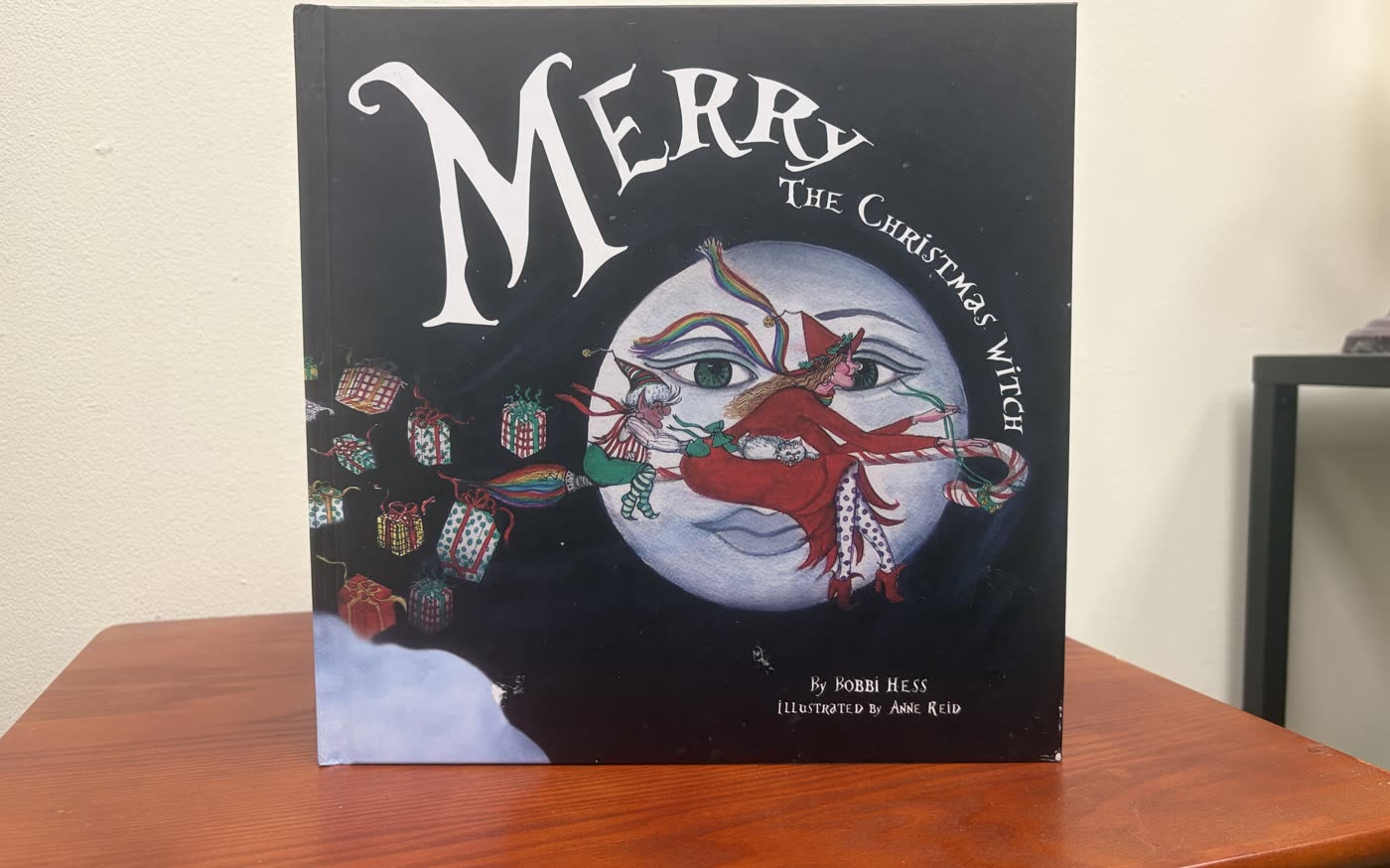 The published picture book Merry the Christmas Witch by Bobbi Hess, illustrated by Anne Reid, standing on a desk.